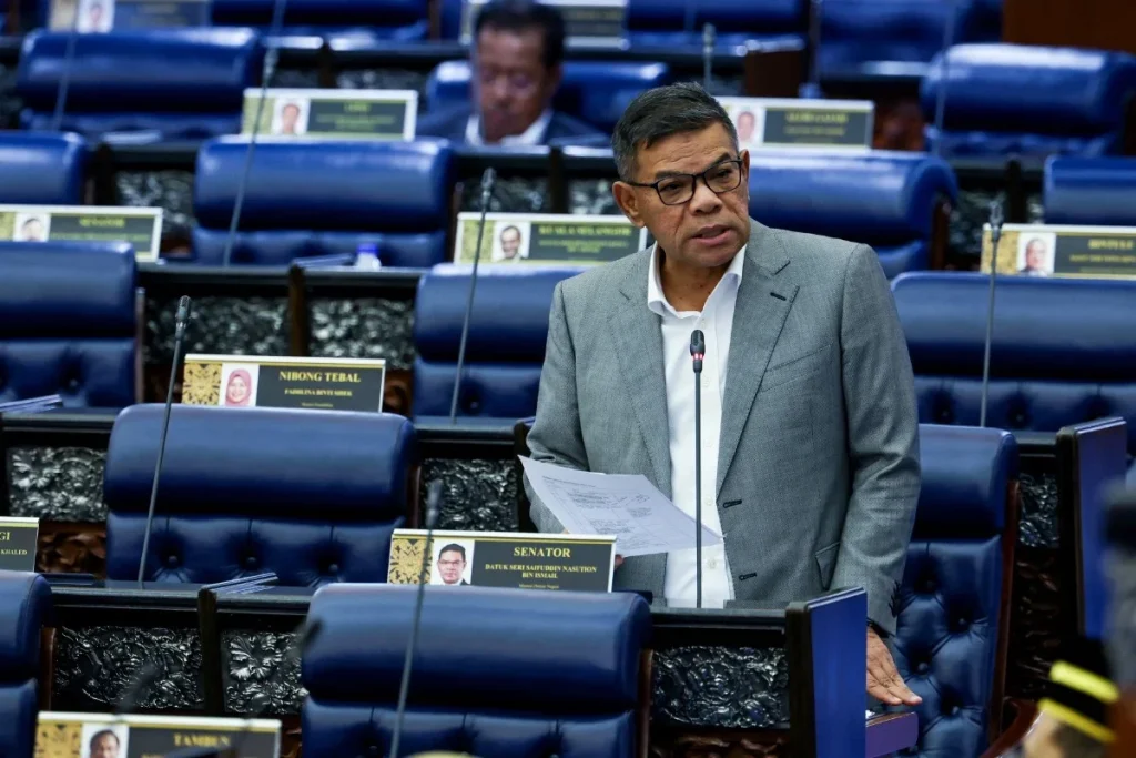 内政部长穿灰色西装在国会发言，站在蓝色座椅之间，手持文件。