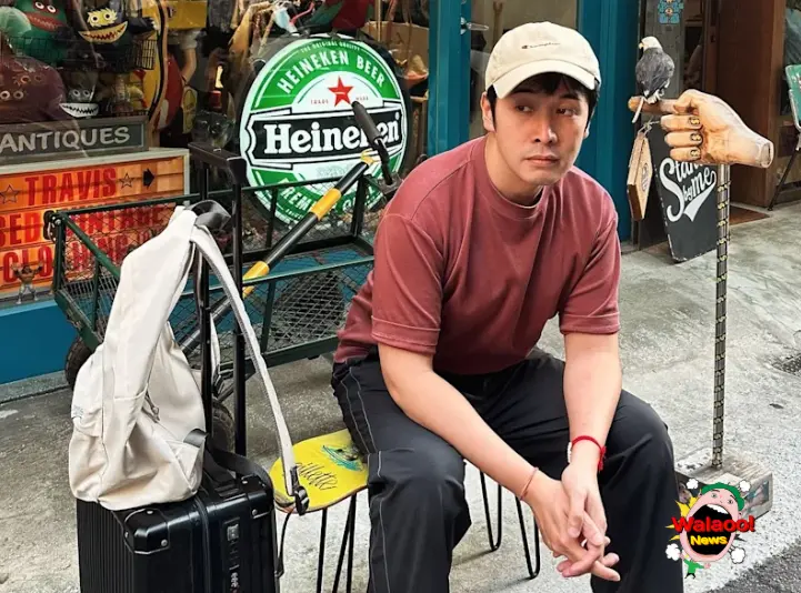 李日朗身穿红色T恤与卡其帽，坐在街边行李箱旁神情憔悴